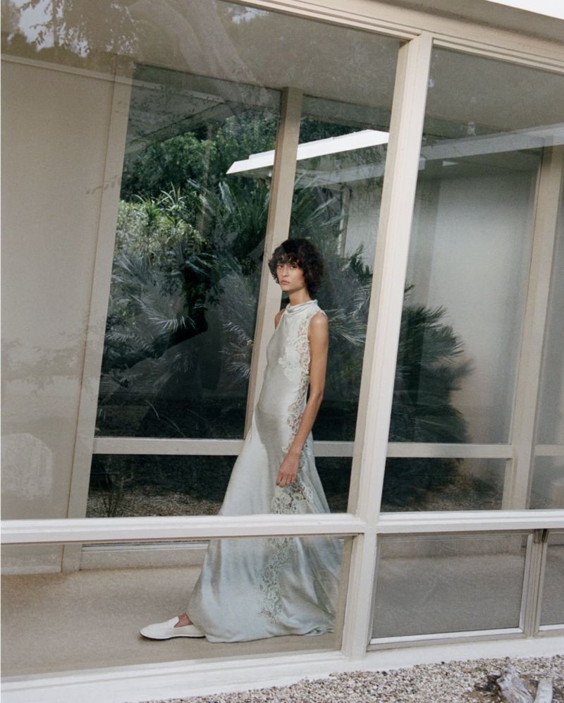 Elegant woman in a sleeveless lace dress poses inside a modern structure with large glass windows, showcasing both fashion and architecture.