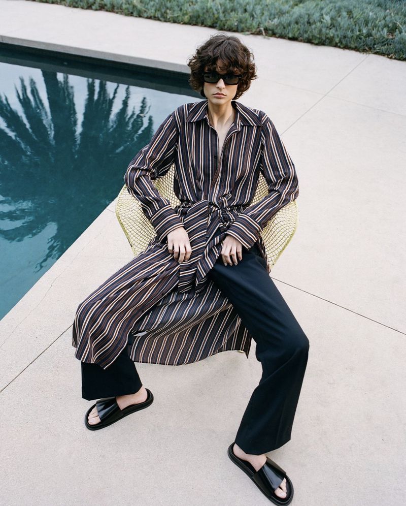 A fashionable adult woman in a striped shirt and black pants poses while seated outdoors in a chair near a swimming pool and foliage.