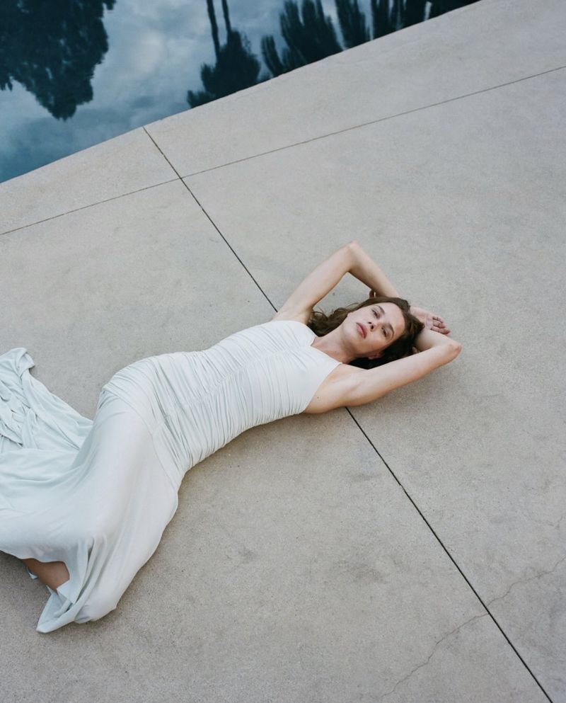 Woman in a white dress lying on a concrete surface next to a swimming pool with a reflection of trees in the water.