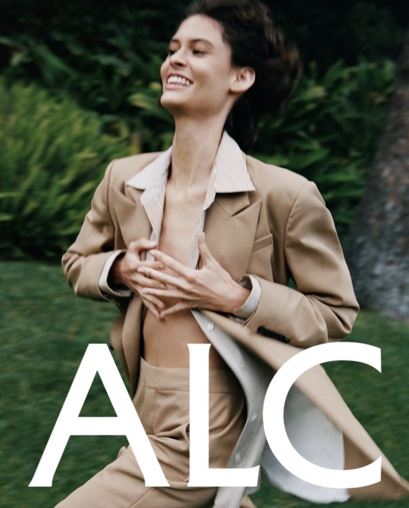 An adult woman smiles and runs in an open, grassy area, wearing a tan suit with an unbuttoned shirt; the brand name ALC is superimposed.