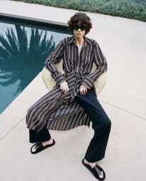 Model wearing dark sunglasses and a striped belted dress seated in a chair by a swimming pool for the A.L.C. collection.