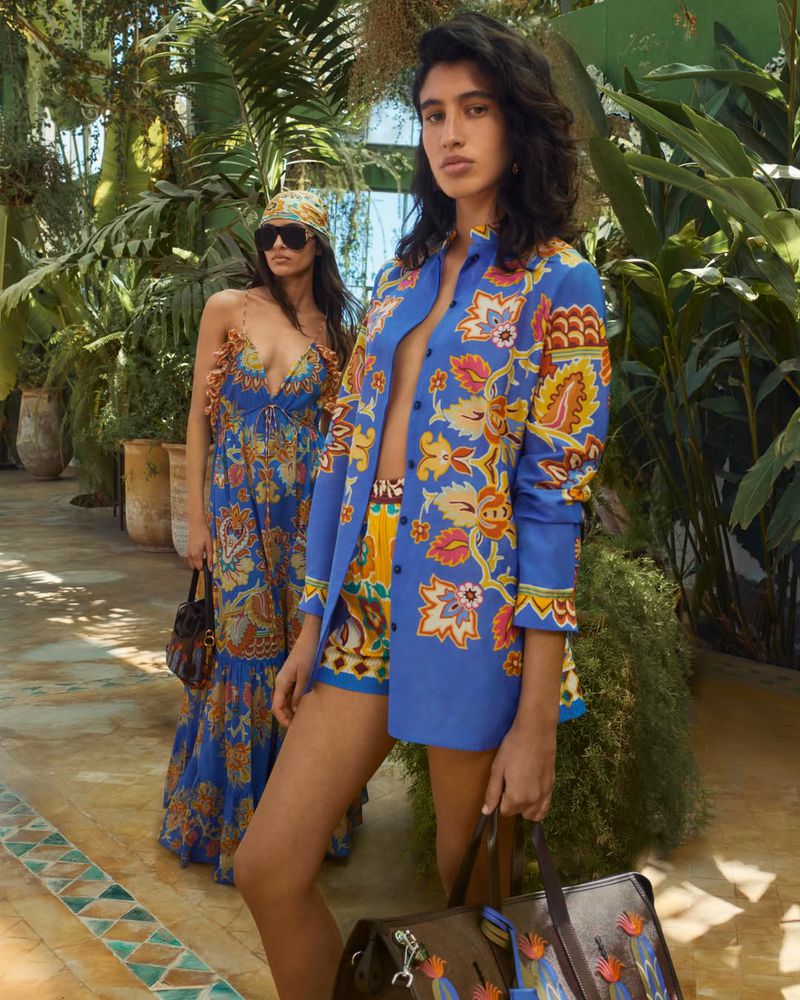Two women model vibrant patterned outfits in a lush, sun-drenched outdoor setting, one in a dress, the other in a jacket and shorts.