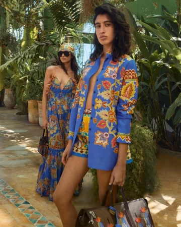 Two women model vibrant patterned outfits in a lush, sun-drenched outdoor setting, one in a dress, the other in a jacket and shorts.