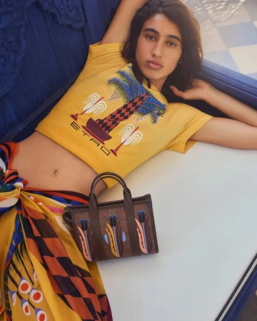 Person poses with a handbag in a fashion setting, wearing an Etro graphic tee and colorful patterned wrap skirt on a blue surface.