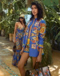 Two female models posing in vibrant patterned resort wear holding leather tote bags amid lush tropical greenhouse foliage.