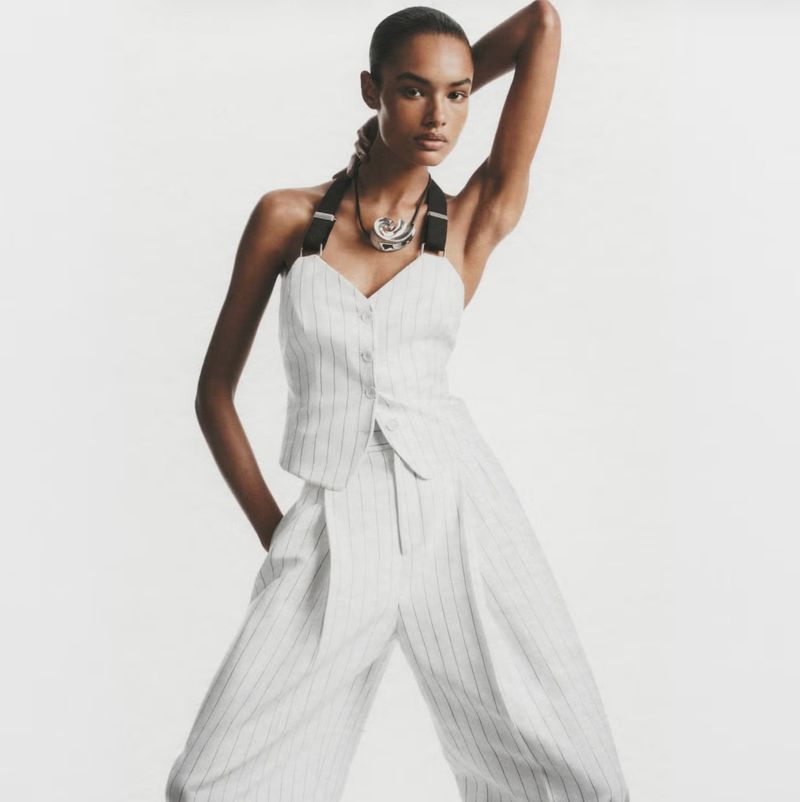 Fashion model poses in a pinstripe top and pants with a unique necklace in a bright, neutral studio setting, creating a chic fashion statement.