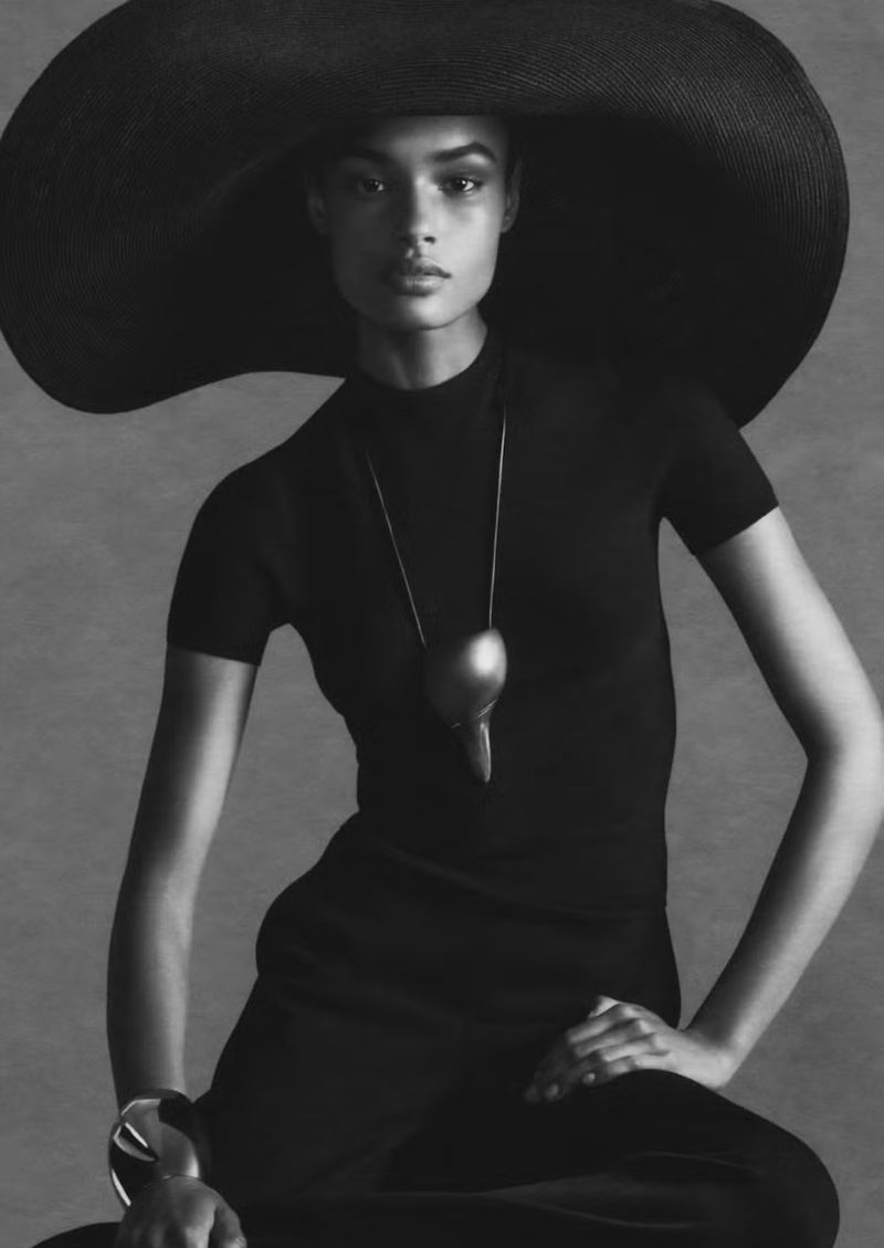 Elegant woman in a black dress and wide-brimmed hat poses in a studio, jewelry necklace, and bracelet, against a grey backdrop.
