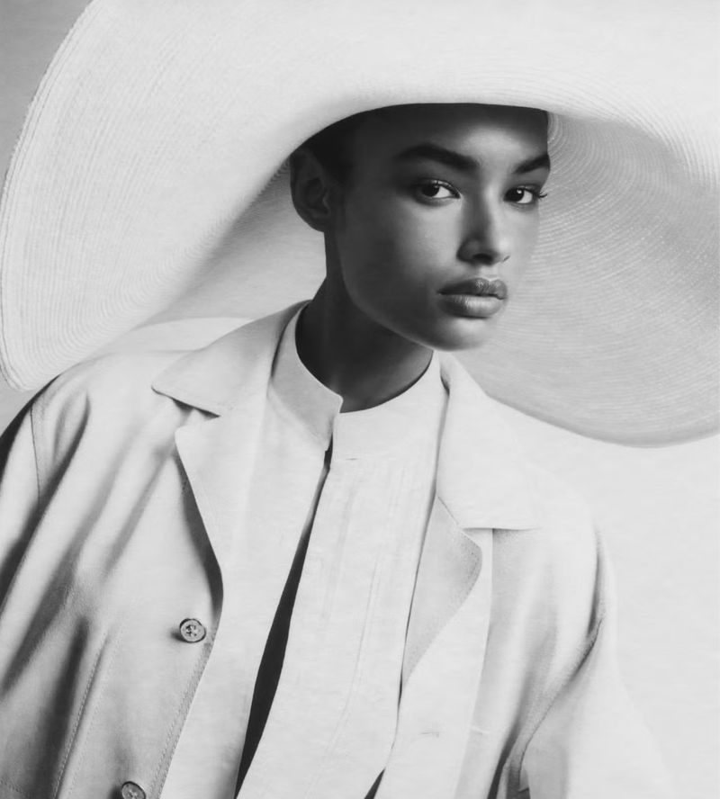 Striking portrait of a Black model in a wide-brimmed hat and a light-colored trench coat against a plain background, fashion photography.