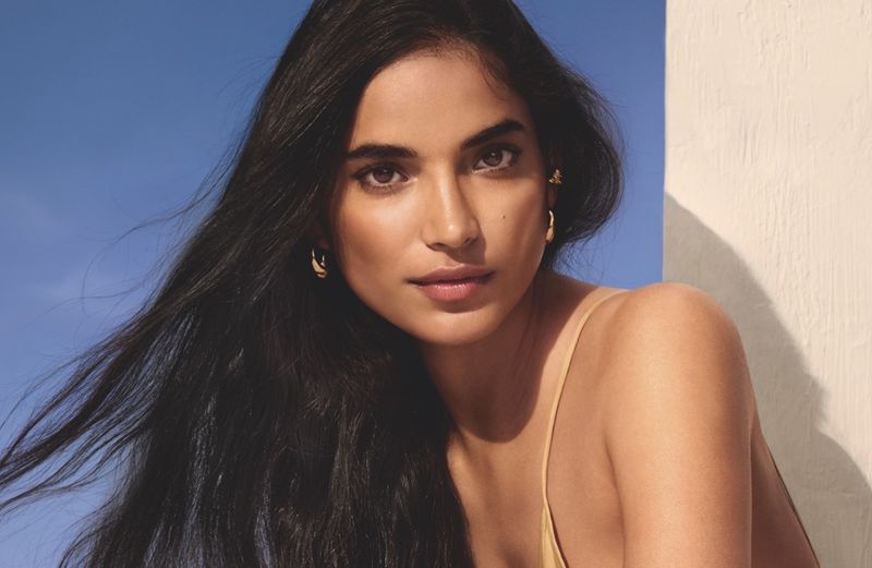 Portrait of a young woman with long, dark hair, golden hoop earrings, and a neutral expression against a light blue and beige backdrop.