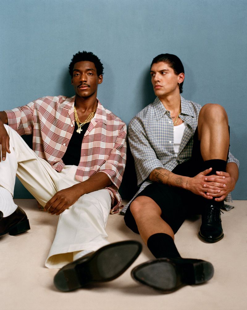 Two stylish men sit indoors. One man wears a red-and-white plaid shirt and cream pants. The other wears a gray plaid shirt and black shorts.