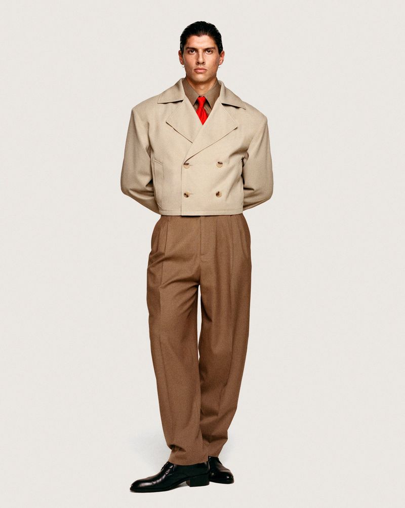 Full studio shot of a male model wearing a beige jacket, brown pants, and black shoes against a white background in a fashion editorial.