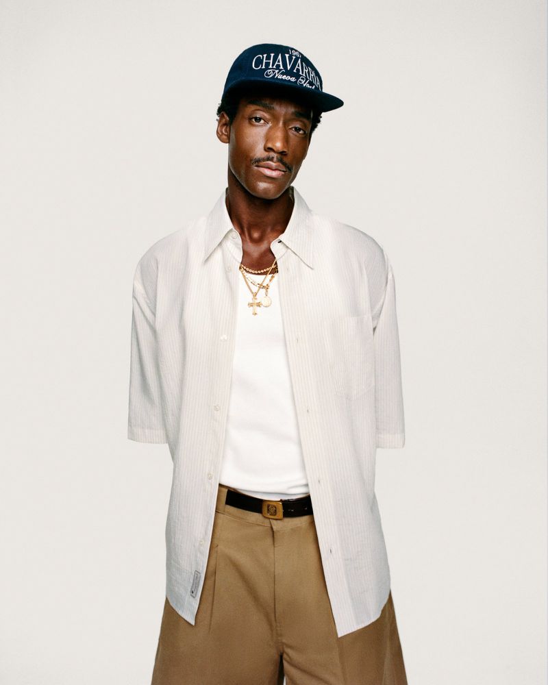 Man in cap and open striped shirt with gold chains, posing in front of neutral background, looking at the camera.