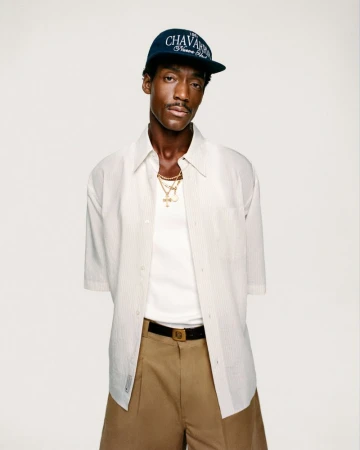 Man in cap and open striped shirt with gold chains, posing in front of neutral background, looking at the camera.