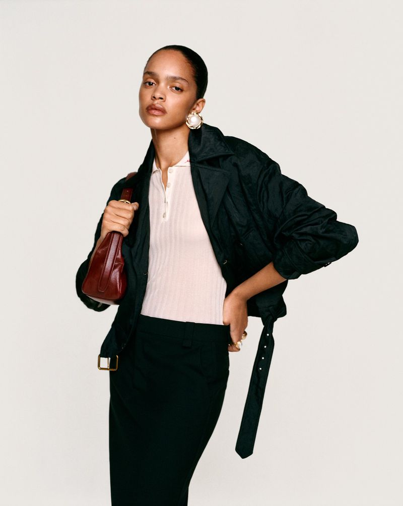 Model wearing a dark jacket, light polo shirt, and holding a red handbag against a seamless white background.