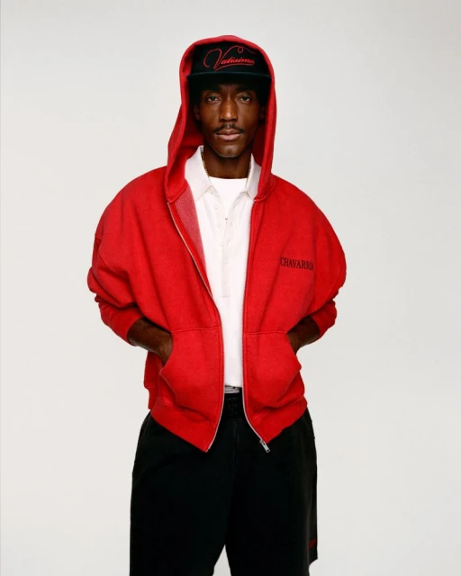 Portrait of a Black man in a red hooded sweatshirt and baseball cap, posing against a white backdrop with hands in his pockets.