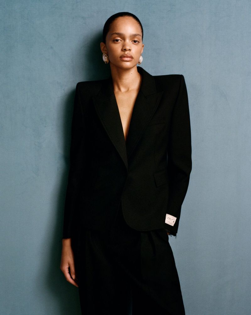 Elegant portrait of a Black woman in a black suit against a textured blue wall, showcasing fashion and beauty.