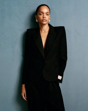 Elegant portrait of a Black woman in a black suit against a textured blue wall, showcasing fashion and beauty.