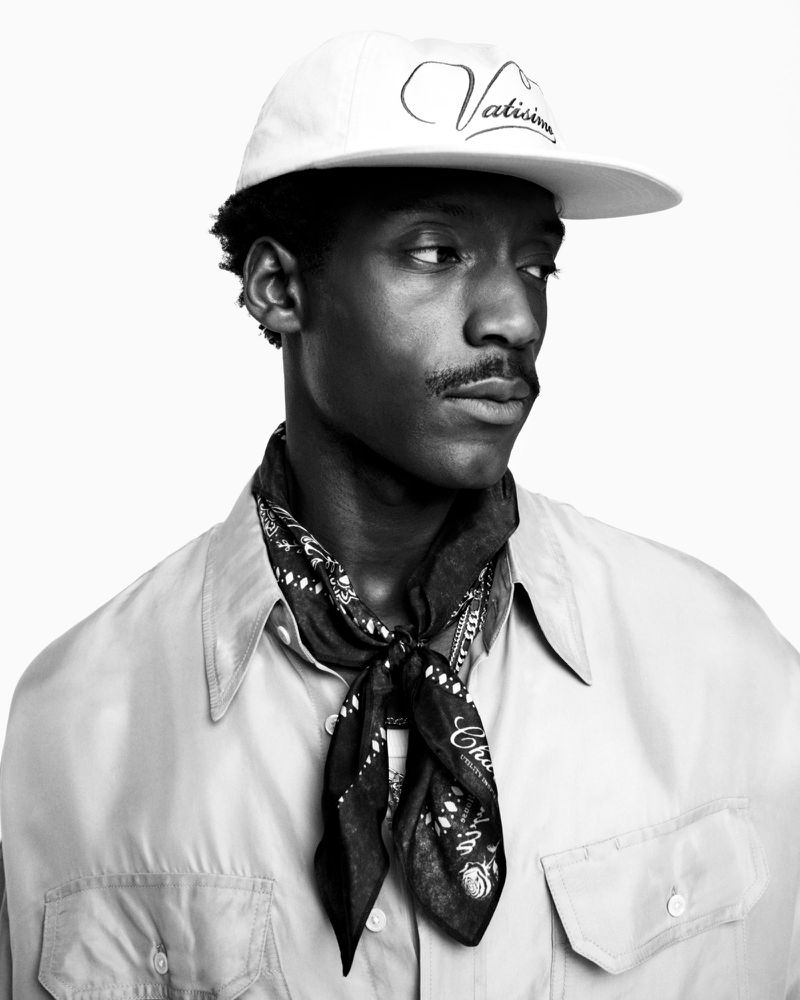 Black and white portrait of a young man with a mustache wearing a baseball cap, shirt, and bandana, against a white backdrop.