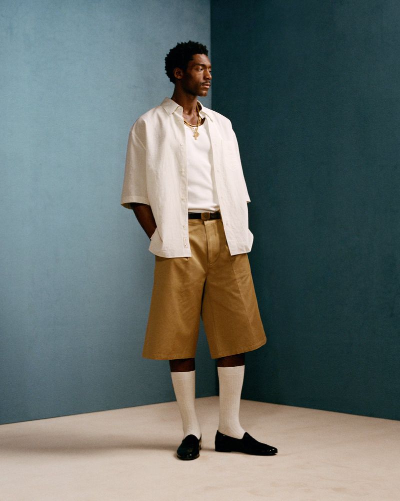 An adult Black man in a fashion pose against a teal and white wall wears an off-white shirt, wide tan shorts, white socks, and black loafers.