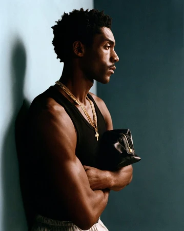 Adult male poses in side profile, wearing a tank top and gold chains, and holding boxing gloves in an indoor environment.