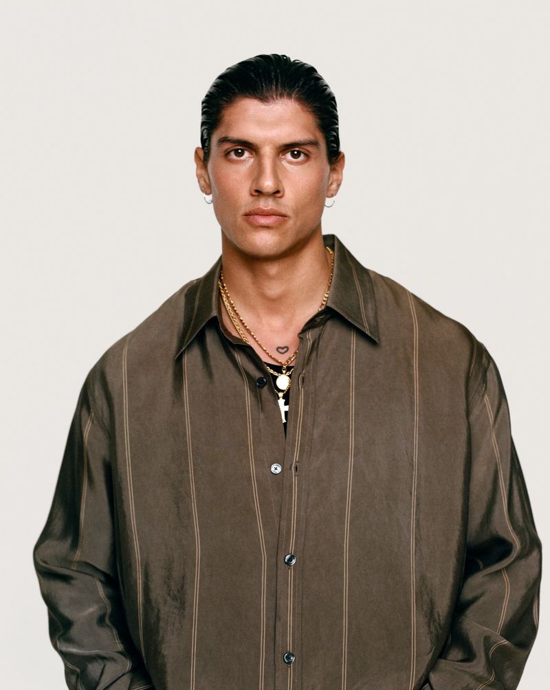 Portrait of a young adult man wearing a button-down shirt and gold jewelry, posing against a neutral background with serious expression.