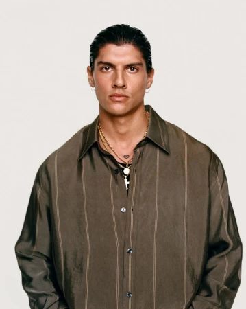Portrait of a young adult man wearing a button-down shirt and gold jewelry, posing against a neutral background with serious expression.