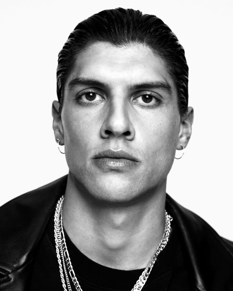 Black and white portrait of adult male with slicked-back hair, wearing a black t-shirt, leather jacket and layered silver chains.