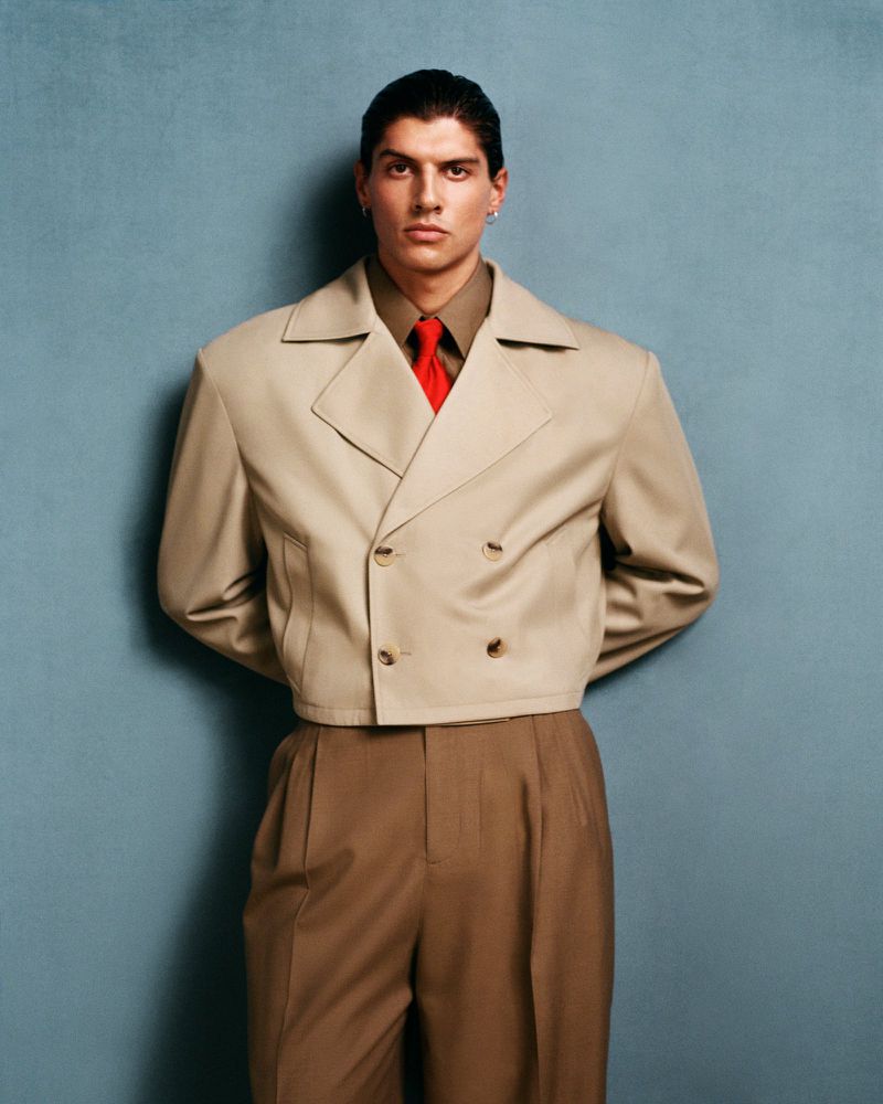 Man wearing a cream double-breasted jacket, olive shirt, and red tie against a textured blue wall.