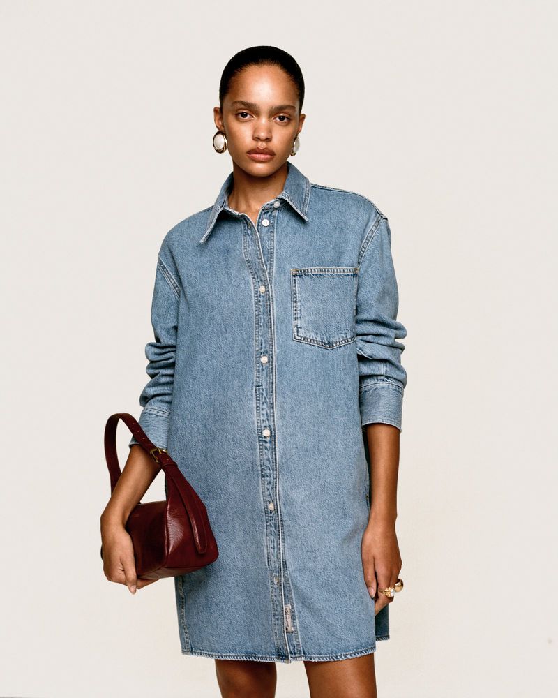 Model with pulled-back hair wearing a blue denim shirt dress and carrying a small deep red leather shoulder bag.