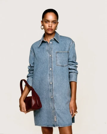 Model with pulled-back hair wearing a blue denim shirt dress and carrying a small deep red leather shoulder bag.