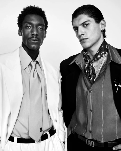 Two men in formal attire pose in a monochromatic portrait. The man on the left wears a white suit and tie, while the other dons a black suit with a bandana.