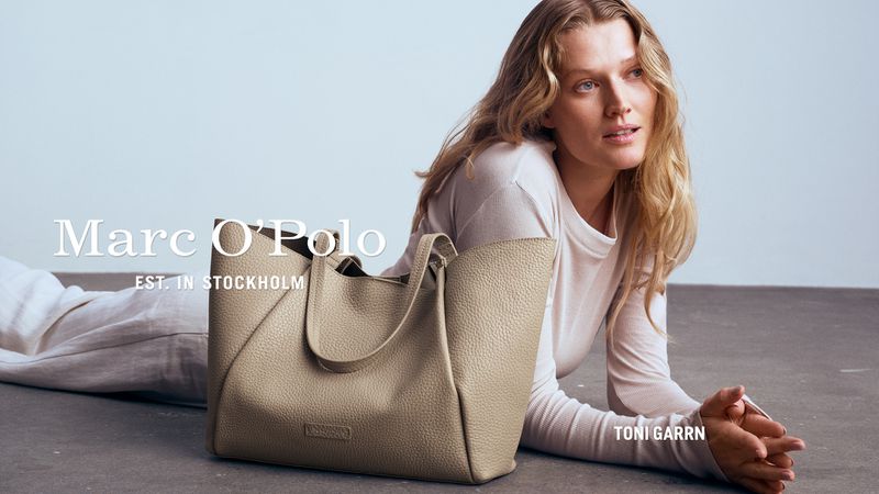 Woman with blonde hair lies on the floor next to a beige leather tote bag in a studio. Fashion advertisement for Marc O'Polo.