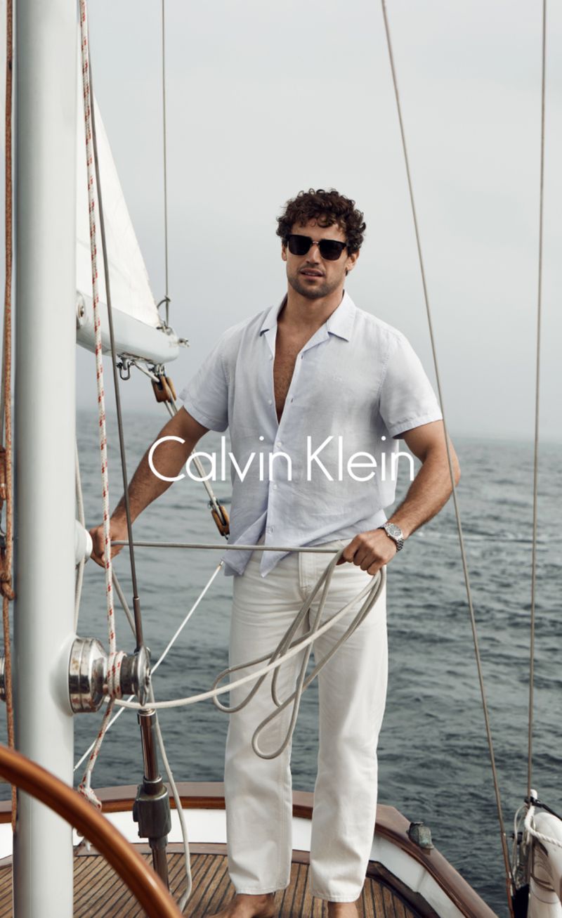 A confident man wearing a linen shirt and cream pants stands on a sailboat, holding a rope, against an ocean and sky backdrop.