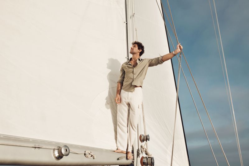 An adult male stands barefoot on the deck of a sailboat wearing an unbuttoned linen shirt and white pants, holding onto the rigging.