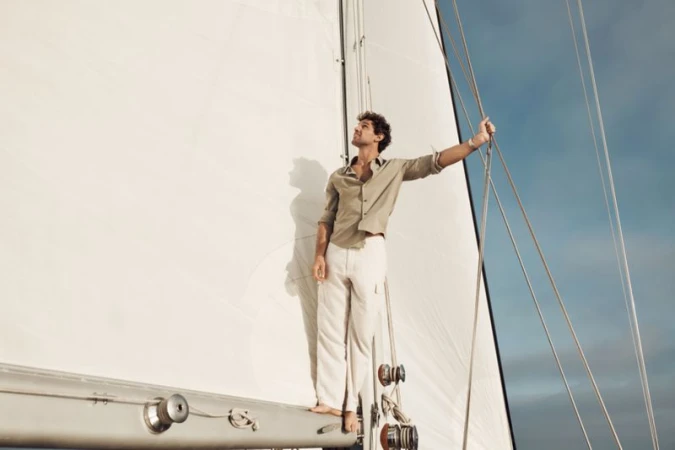 An adult male stands barefoot on the deck of a sailboat wearing an unbuttoned linen shirt and white pants, holding onto the rigging.