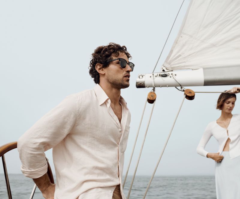 Two people on a sailboat on the ocean. A man in sunglasses and a linen shirt stands near a woman in a white sweater.