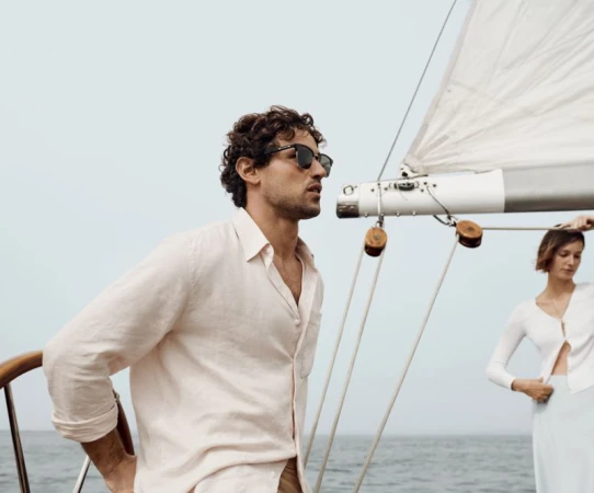Two people on a sailboat on the ocean. A man in sunglasses and a linen shirt stands near a woman in a white sweater.