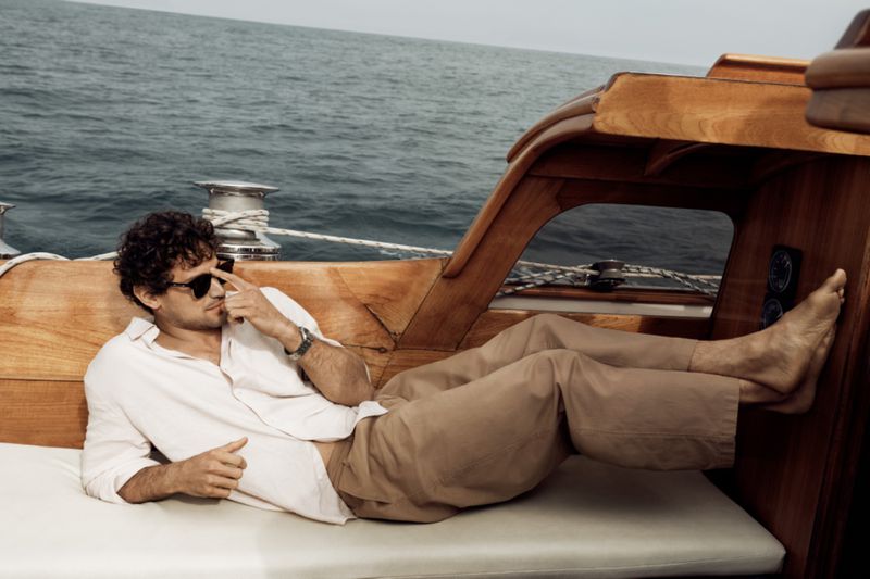An adult male wearing sunglasses and a white shirt is reclining on a boat with a clear view of the ocean in the background.
