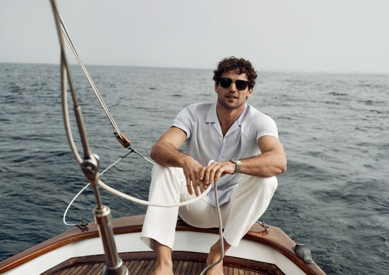 Adult male wearing sunglasses sits on the bow of a wooden boat sailing on the ocean under a cloudy sky in a medium shot.