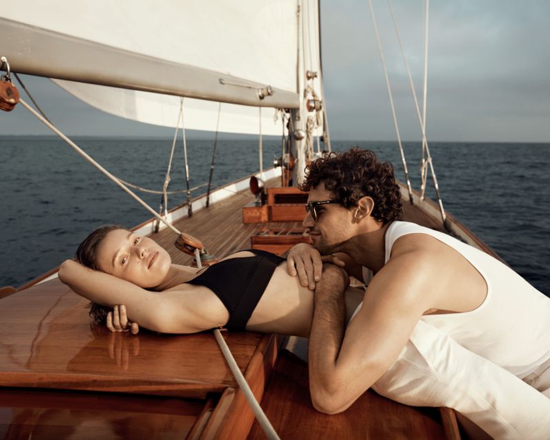 A couple relaxes on a sailboat deck on a sunny day. The woman in a black bikini lies on the wooden deck, while the man sits beside her.