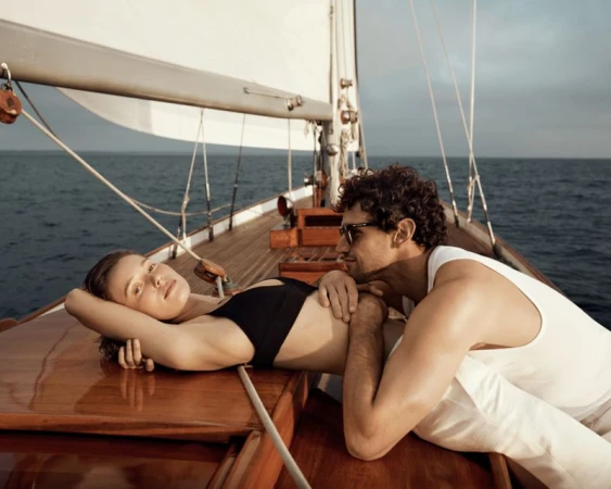 A couple relaxes on a sailboat deck on a sunny day. The woman in a black bikini lies on the wooden deck, while the man sits beside her.
