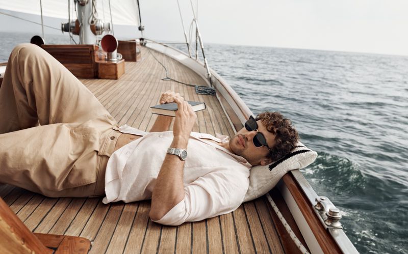 Adult male with sunglasses laying on a sailboat, reading a book, wearing beige linen outfit in a medium shot with ocean background.