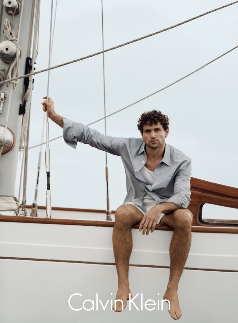 A bare-chested man with curly hair sits on a white sailboat, wearing an unbuttoned light blue shirt, promoting Calvin Klein.