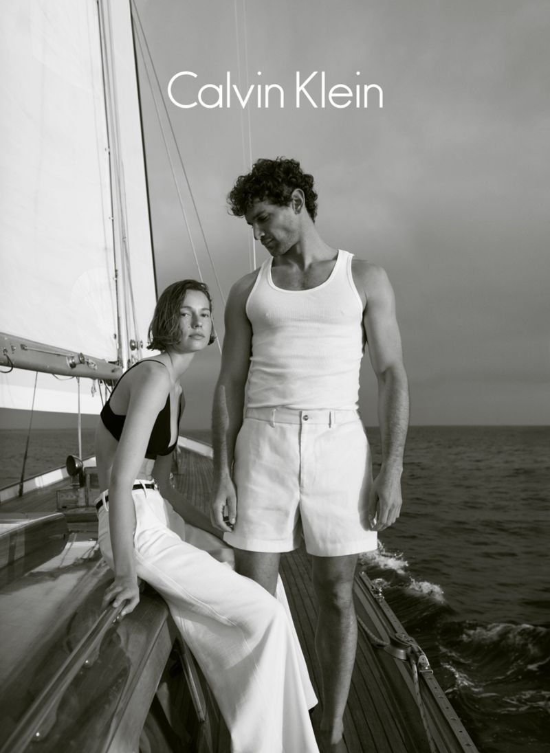 Black and white fashion shot on a sailboat features a couple, with the woman sitting and the man standing, both in white clothing.