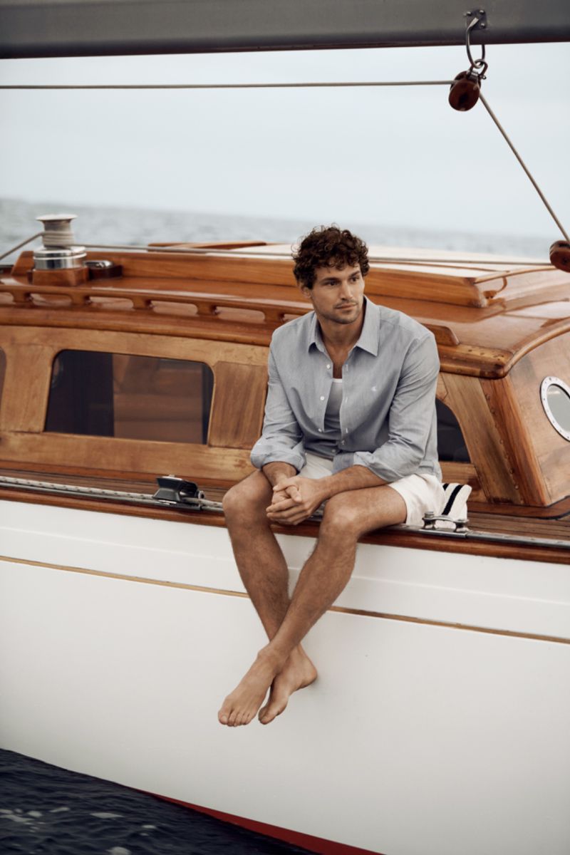 Man sits barefoot on a wooden sailboat deck wearing an unbuttoned shirt, shorts, and looking out towards the calm blue sea.