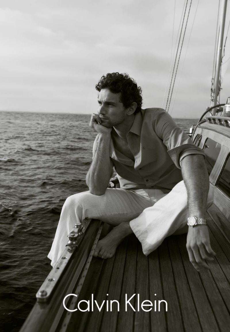Black and white portrait of a man sitting on a sailboat wearing casual linen clothes, on the water, with the Calvin Klein logo visible.