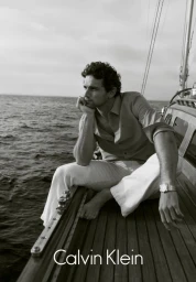 Model Giacomo Cavalli sitting barefoot on a wooden sailboat deck overlooking the ocean for Calvin Klein Menswear.