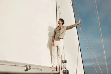 Model Giacomo Cavalli wearing a light shirt and trousers standing on a sailboat deck holding rigging lines