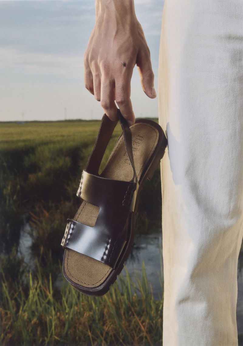 Close-up of a hand holding a brown leather sandal against a blurred outdoor background of green grass and blue sky.