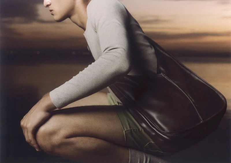 Adult male squats with a brown leather bag at his side, soft sunset background, soft lighting, fashion or product shot, lifestyle.
